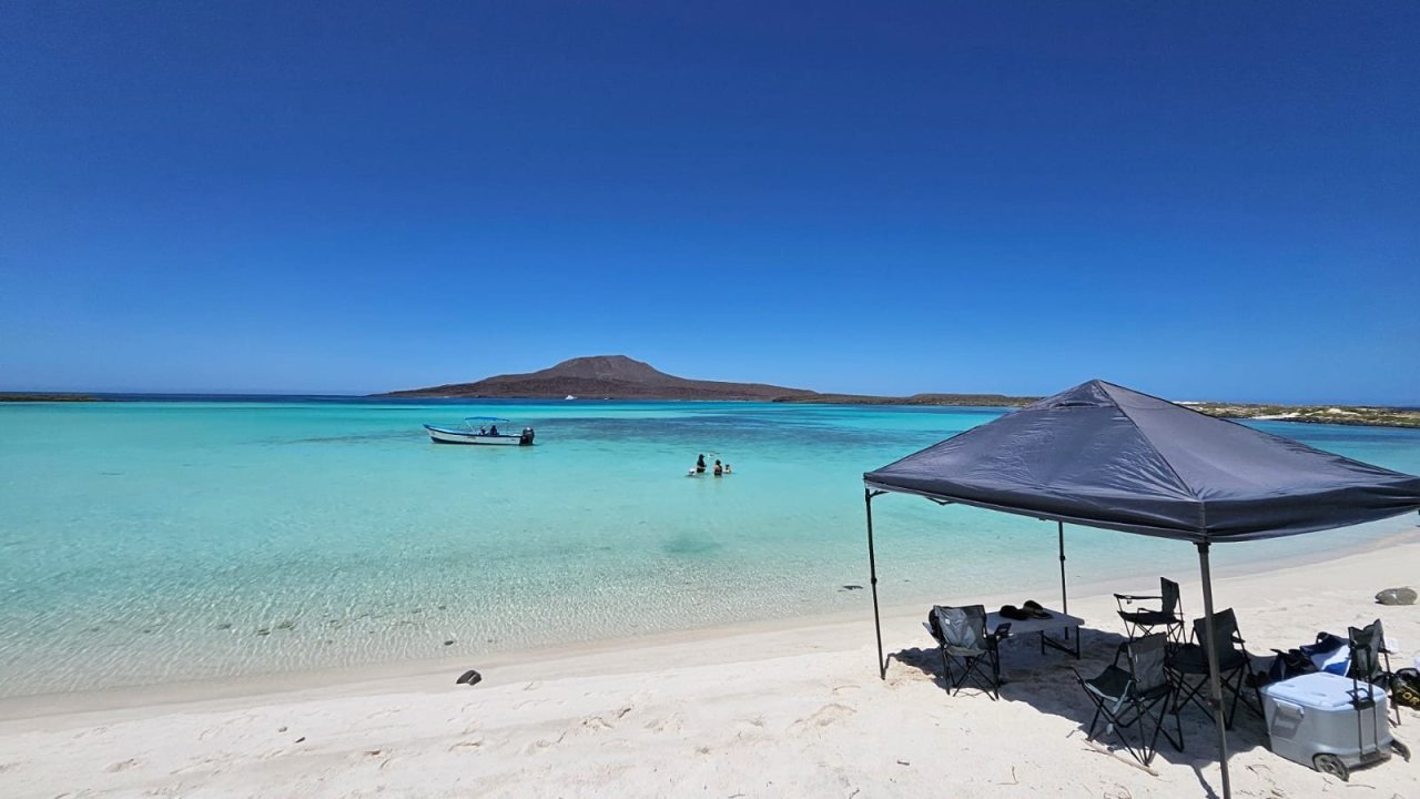 Carpa de sombra, sillas e hielera montadas en la playa de arena blanca de Isla Coronado, con la panga Loreto Star fondeada en agua turquesa