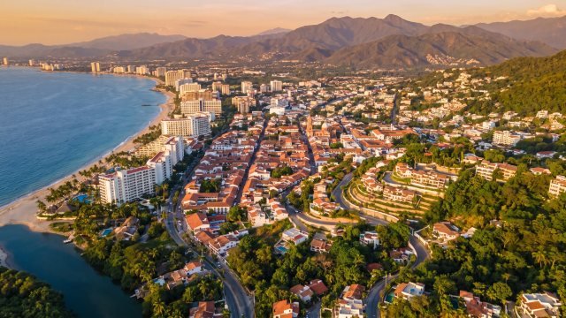 Vista aérea de Puerto Vallarta al atardecer con la franja hotelera, el centro histórico, las colonias en la ladera y la Sierra Madre al fondo
