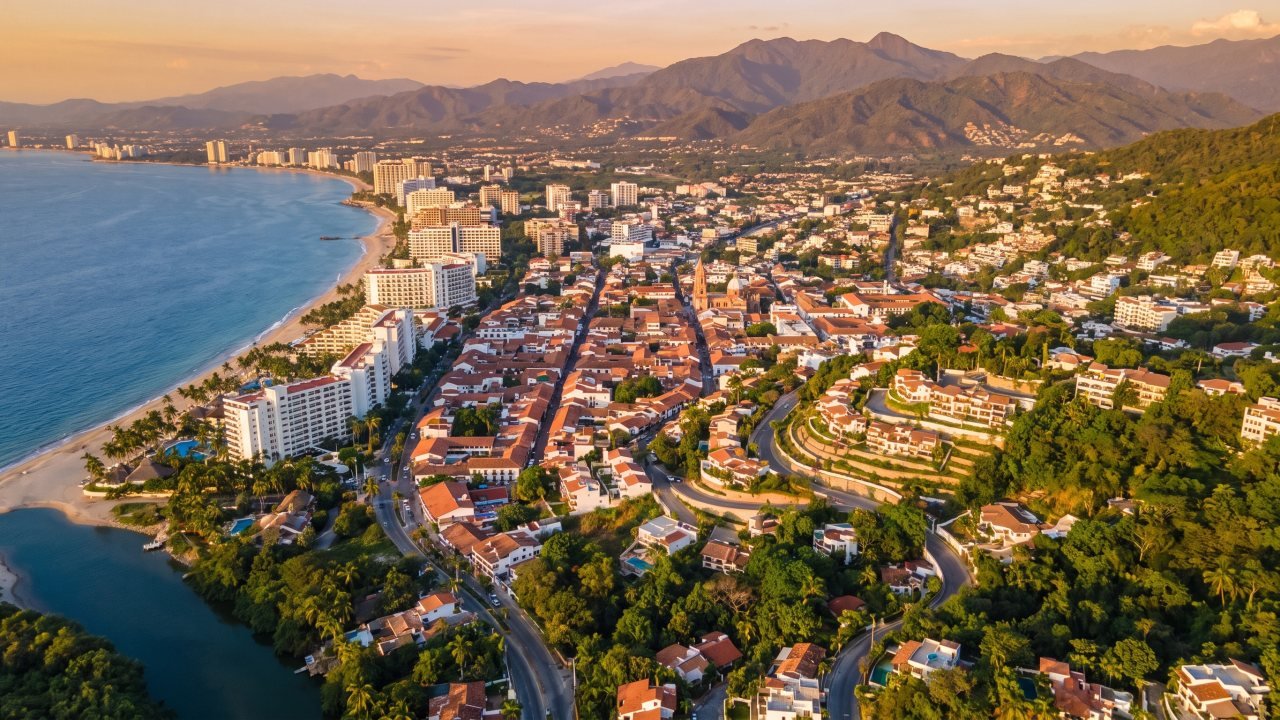Vista aérea de Puerto Vallarta al atardecer con la franja hotelera, el centro histórico, las colonias en la ladera y la Sierra Madre al fondo